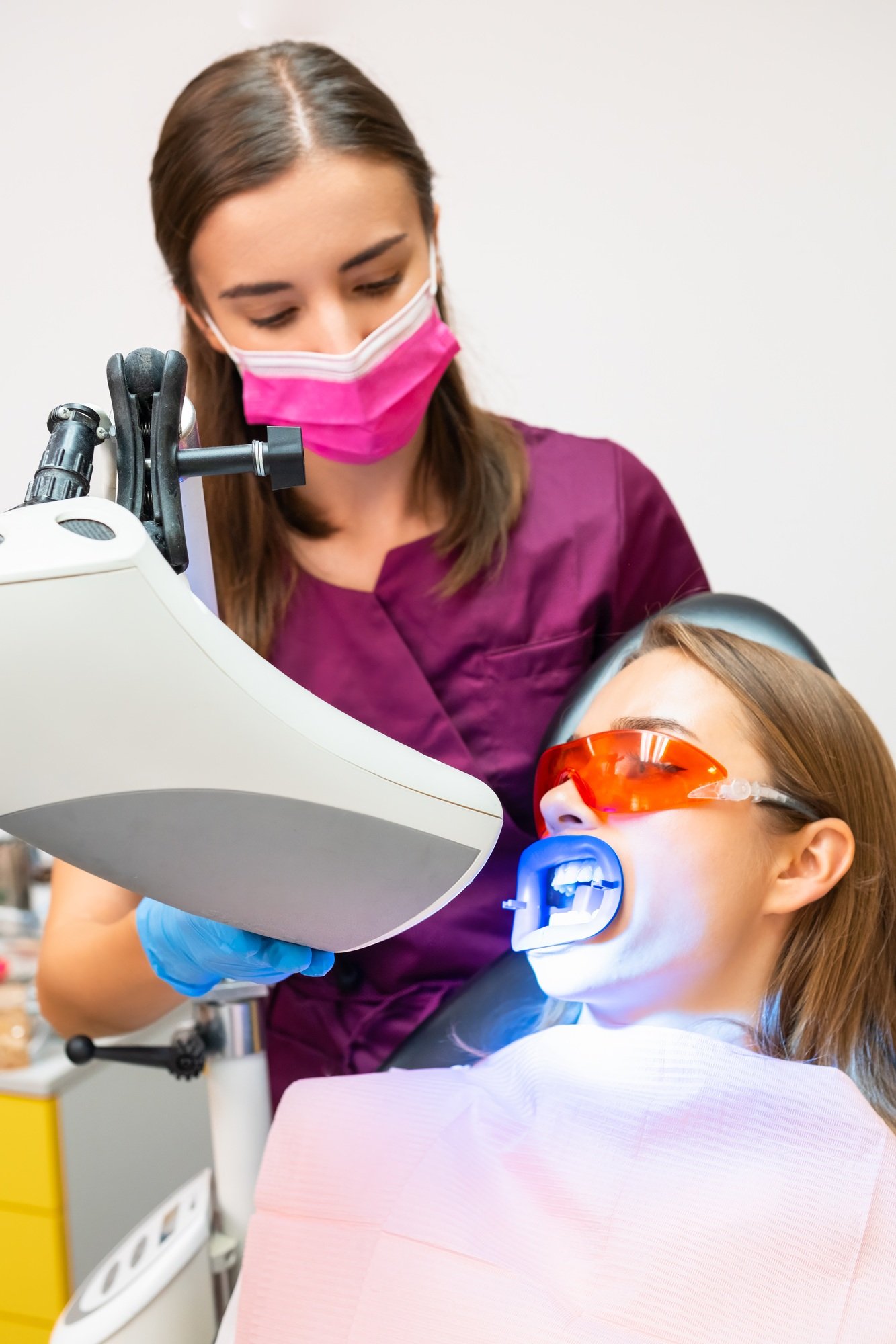 Young dentist in face mask performs whitening of teeth using UV lamp for woman at dentist clinic